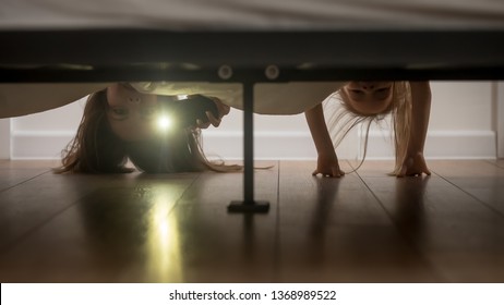 Mother and daughter, babysitter and kid girl afraid of monsters hold smartphone shine a flashlight under the bed looking on floor check ghosts, older younger sisters playing having fun at home concept
