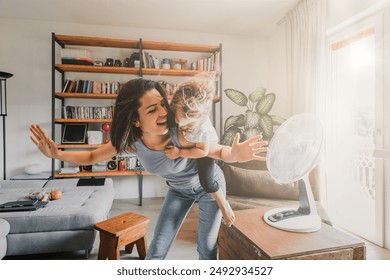 Mother and daughter beat the summer heat at home, playing with an electric fan, creating joyful memories in their cozy living room filled with warm natural light, embodying family togetherness - Powered by Shutterstock