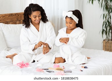Mother Daughter Beauty Treatments. Caring Black Mom Teaching Little Girl Self-Care At Home, Sitting In Bathrobes On Bed, Applying Body Lotion On Hands, Different Makeup Cosmetics Lying Around - Powered by Shutterstock