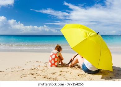 Mother And Daughter With Big Yellow Umbrella Hiding From Sun At Tropical Beach