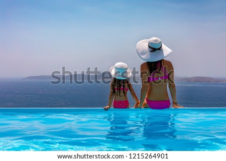 Mother and daughter in bikini are sitting at the pool edge and enjoying the view to the sea during their summer vacation time