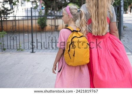Mother and daughter, carrying yellow backpack, head to school, their backs in view. 