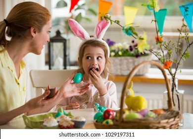 Mother And Daughter Celebrating Easter, Eating Chocolate Eggs. Happy Family Holiday. Cute Little Girl With Funny Face In Bunny Ears Laughing, Smiling And Having Fun.