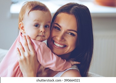 Mother And Daughter. Cheerful Beautiful Young Woman Holding Baby Girl In Her Hands And Looking At Camera With Smile While Sitting On The Couch At Home