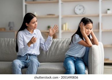 Mother daughter conflict. Young korean mother trying to speak to her upset daugter, sitting on sofa at home. Offended asian girl covering her ears, not wishing to listen to parent
