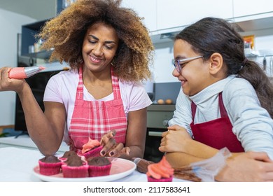 Mother And Daughter Cooking Pastries In The Kitchen, Latin Adult Woman Decorating Cupcakes With Sac A Poche Filled With Pink Sweety Cream, Genuine Moment Of Parent Chilld Bonding