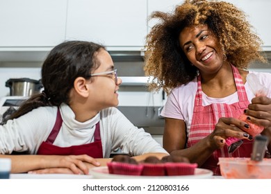 Mother And Daughter Cooking Pastries In The Kitchen, Latin Adult Woman Decorating Cupcakes With Sac A Poche Filled With Pink Sweety Cream, Genuine Moment Of Parent Chilld Bonding