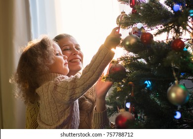 Mother And Daughter Decorate A Christmas Tree Against The Window With A Setting Sun And Bright Sunlight