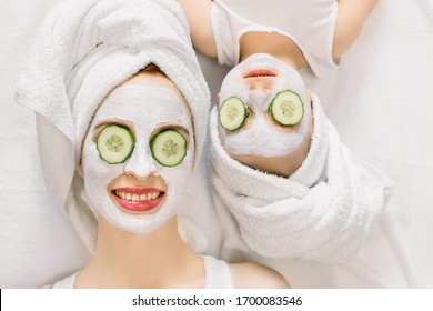 Mother And Daughter Doing Funny Spa Procedures After Bath. They Are In White Bath Towels With White Facial Mud Mask On Faces And Slices Of Fresh Cucumber On Their Eyes. Family Spa, Mothers Day