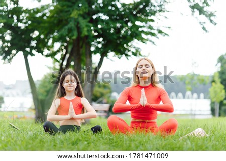 Mother and daughter doing yoga exercises on grass in the park at the day time. People having fun outdoors. Concept of friendly family and of summer vacation.