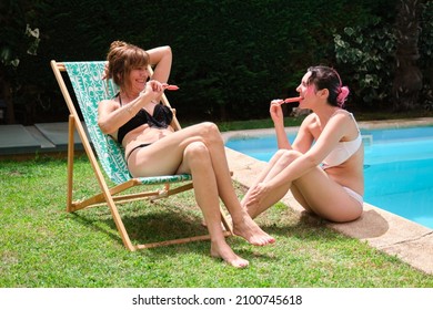 Mother and daughter eating a strawberry popsicle sitting near by a pool.