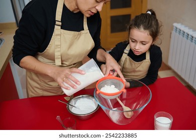 A mother and daughter enjoy a baking session, sifting flour together in a homely kitchen setting. The little girl attentively watches, wearing matching aprons, fostering a warm family atmosphere. - Powered by Shutterstock