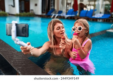 Mother and daughter enjoy ice cream in pool, selfie stick in hand, capturing smiles beside blue water. Mom and girl in swimwear share sweet moment, taking summer memories with smartphone. - Powered by Shutterstock