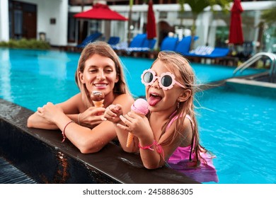 Mother and daughter enjoy poolside ice cream on sunny day. Child in pink swimsuit, goggles bites cone, woman smiles. Water splashes near sun loungers, umbrellas. Family pool fun, summer treat bonding. - Powered by Shutterstock