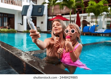 Mother and daughter enjoy summer day in pool with selfie stick, capturing moment while eating ice cream. Woman in swimsuit with girl in pink, smiles at camera, leisure fun, family vacation bonding. - Powered by Shutterstock