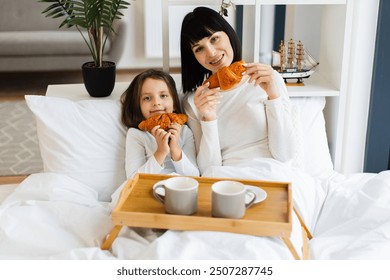 Mother and daughter enjoying breakfast in bed with hot drinks and fresh croissants. Demonstrates family bonding, morning routine, and happiness. - Powered by Shutterstock