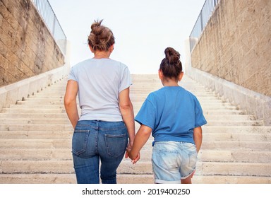 Mother And Daughter Going Up The Stone Stairs. A Middle-aged Woman And Kid Girl Holding Each Other Hands And Wearing Blue T-shirts Back View. Motherhood Concept