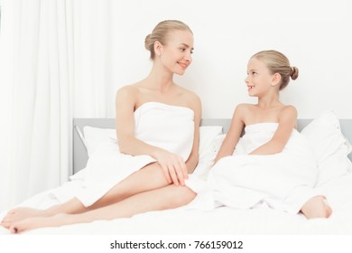 Mother And Daughter Had A Day Of Spa. They Have Fun In White Bath Towels. They Sit On The Bed And Look At Each Other.