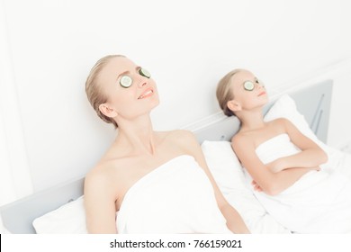 Mother And Daughter Had A Day Of Spa. They Are In White Bath Towels With Slices Of Cucumber On Their Eyes.