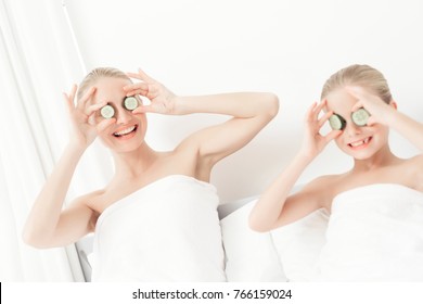 Mother And Daughter Had A Day Of Spa. They Are In White Bath Towels With Slices Of Cucumber On Their Eyes.