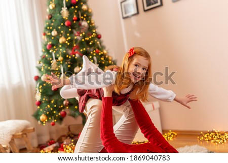 Mother and daughter having fun playing at home on Christmas day, mother lying on the floor by the Christmas tree and lifting her little daughter