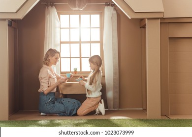 Mother And Daughter Having Tea Party In Cardboard House Against Window