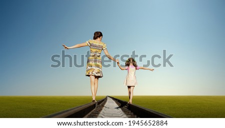 mother with daughter hold hands and walk on railway tracks, on green field and blue sky background