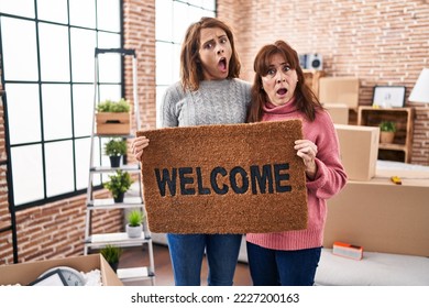 Mother and daughter holding welcome doormat in shock face, looking skeptical and sarcastic, surprised with open mouth  - Shutterstock ID 2227200163