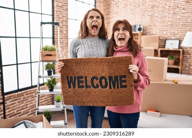 Mother and daughter holding welcome doormat angry and mad screaming frustrated and furious, shouting with anger looking up.  - Shutterstock ID 2309316289