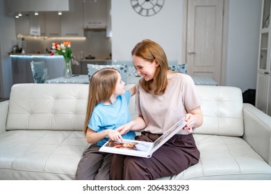 Mother And Daughter At Home On The Couch Leafing Through And Looking A Book With Photos From A Family Photo Shoot With A Newborn Baby. Memory Of Important Moments Of Life In The Photo Album.