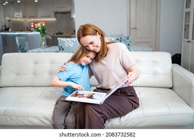 Mother And Daughter At Home On The Couch Leafing Through And Looking A Book With Photos From A Family Photo Shoot With A Newborn Baby. Memory Of Important Moments Of Life In The Photo Album.