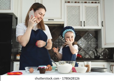 A Mother And Daughter In Identical Aprons And Chef's Hats Are Cooking In The Kitchen, Smiling And Looking At The Camera. Mom's On The Phone.