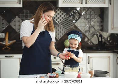 A Mother And Daughter In Identical Aprons And Chef's Hats Are Cooking In The Kitchen, Smiling And Looking At The Camera. Mom's On The Phone.