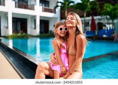 Mother and daughter laugh by luxury resort pool in tropics. Family enjoy sunny vacation, swimwear fashion, quality time together. Mom hugs girl, tropical holiday relax, leisure lifestyle, happiness. - Powered by Shutterstock