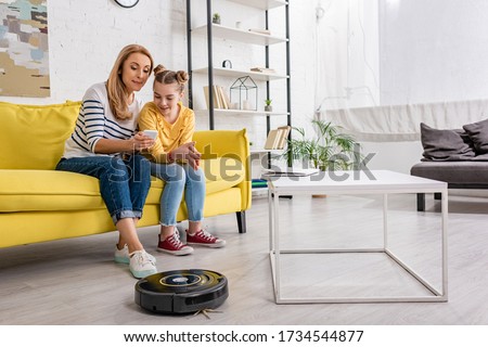 Mother and daughter looking at smartphone on sofa near coffee table with laptop and robotic vacuum cleaner on floor in living room Stock photo © 