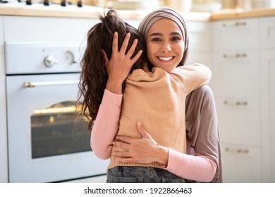 Mother Daughter Love. Beautiful Muslim Lady In Hijab Embracing Her Little Child In Kitchen Interior, Small Girl Hugging Her Happy Islamic Mom At Home, Greeting With Mother's Day, Free Space