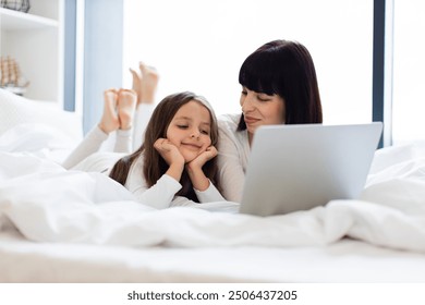 Mother and daughter lying in bed using laptop. Child resting head on hands smiling at screen. Family bonding moment with technology in cozy bedroom. Happy and relaxed atmosphere. - Powered by Shutterstock