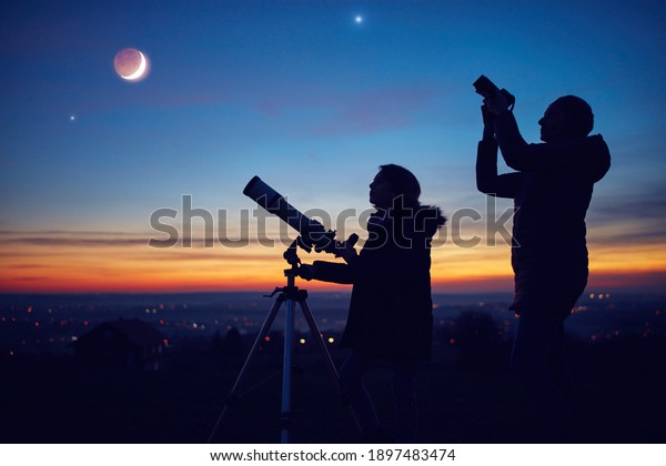 Mother and daughter observing\
stars, planets, Moon and night sky with astronomical\
telescope.