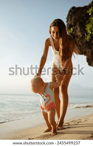 Mother and daughter on the coast. Mom teaches daughter to make first steps on the sand near the sea. Portrait of happy loving mother and her baby outdoors during summer beach vacation