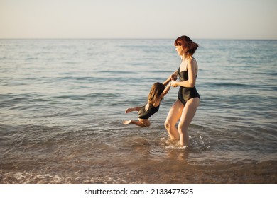 Mother And Daughter On Sea Rest. Family Are On Sea Beach. Mom And Daughter In Black Swimwear Playing On Sea Water. Woman With Girl Swimming And Splashing In Sea.