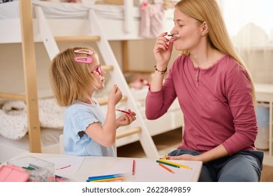 mother and daughter playing with colored pencils pretending to have mustaches. brightly decorated children's room captures playful, creative family bonding moment during indoor fun activities