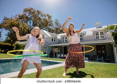 Mother And Daughter Playing With Hula Hoop In Their Backyard On A Summer Day. Happy Young Family Having Fun With Hula Hoop Outdoors.