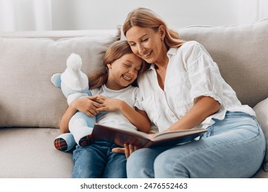 Mother and daughter reading a book together on a cozy living room couch, sharing quality family time - Powered by Shutterstock