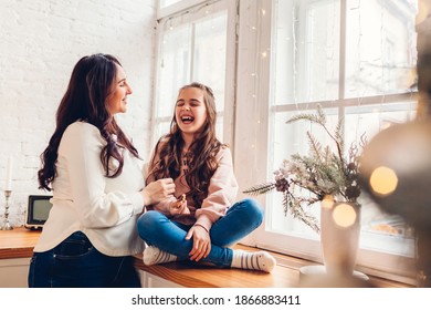 Mother And Daughter Relaxing On Kitchen Window Sill By Decorated Christmas Tree. Family Having Fun Together At Home