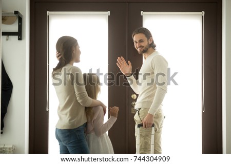 Mother and daughter seeing off father leaving for business trip, mom with kid girl saying goodbye to dad waving hand standing at home door holding travel case ready to go, family separation, good bye 