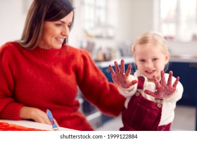 Mother With Daughter Showing Messy Hands At Home Doing Craft And Painting Picture In Kitchen