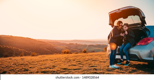 Mother and daughter sitting in a car trunk on a mountain and using smartphone - Powered by Shutterstock