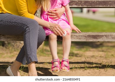 Mother And Daughter Sitting On Bench. Parent Hand Using White Cotton Pad On Toddler Girl Abrasion Knee Skin After Fell Down In Summer Day. Giving First Aid For Child. Closeup. Front View.