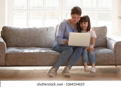 Mother and daughter sitting on couch spends time in living room at home having fun together using computer apps, browsing internet, watching cartoon online, mom teach kid electronic device use concept