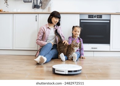 Mother and daughter sitting on kitchen floor playing with pet cat while robotic vacuum cleaner operates. Bright and cozy kitchen setting promoting family bonding, love for pets. - Powered by Shutterstock